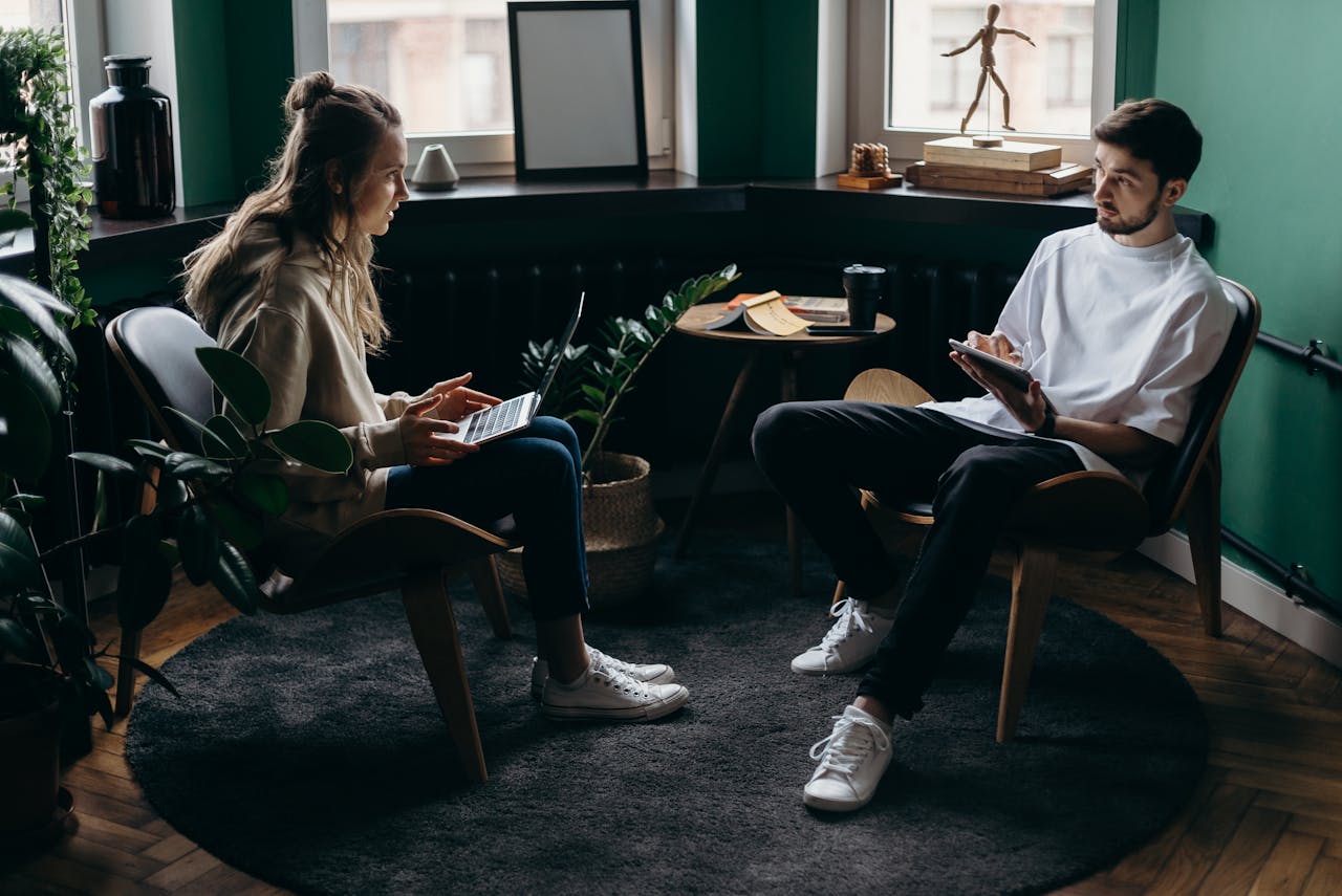 gallery-3 A man and woman engaged in conversation at a stylish, plant-filled home office with modern furniture and digital devices.